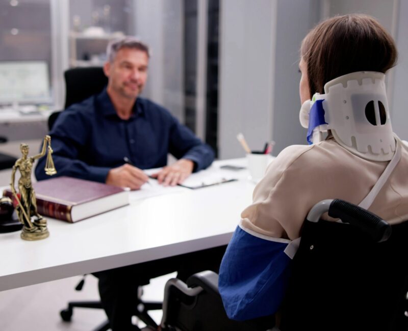 Injured woman in a wheelchair wearing a neck brace and arm sling consulting with a car accident lawyer in a modern legal office.