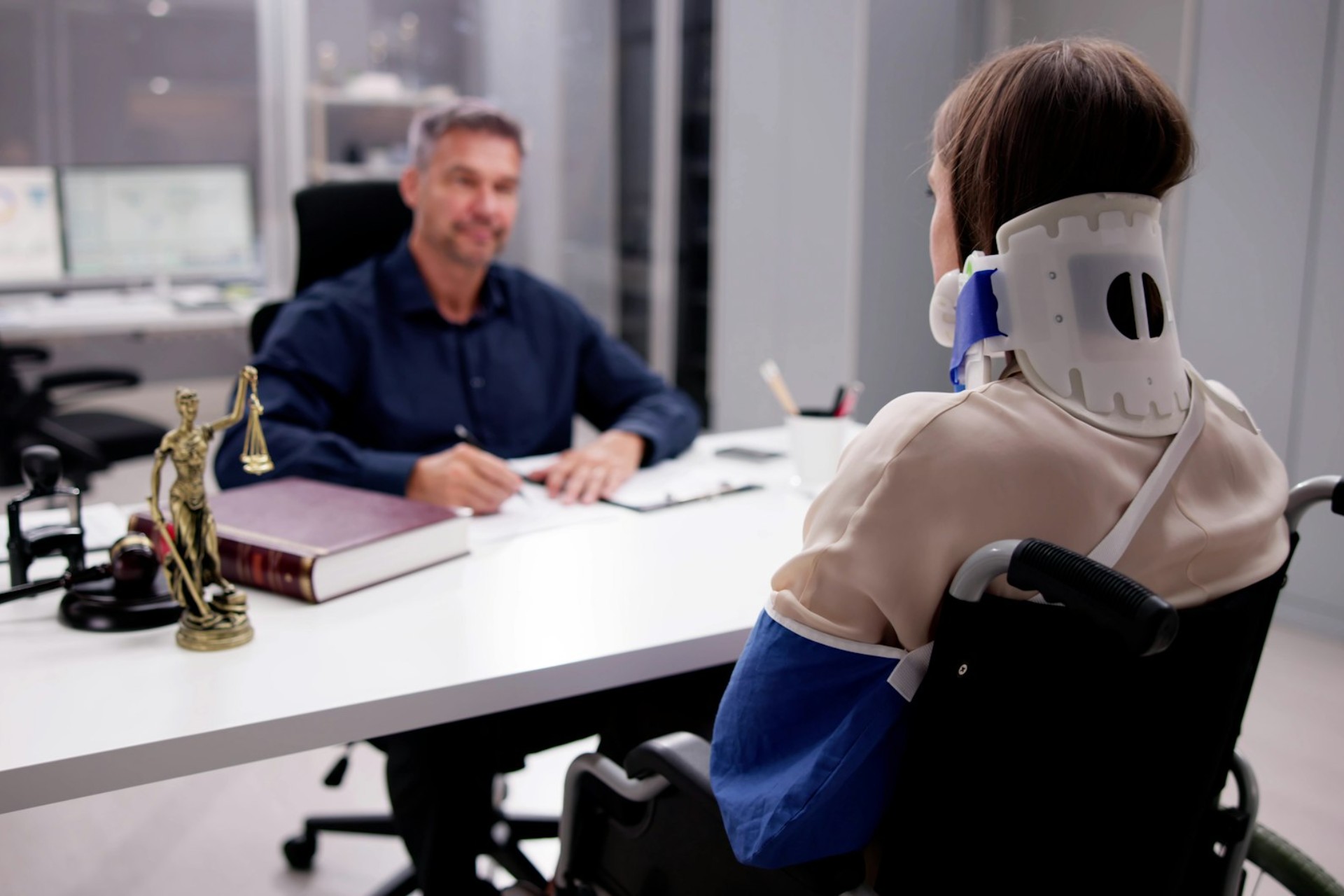 Injured woman in a wheelchair wearing a neck brace and arm sling consulting with a car accident lawyer in a modern legal office.