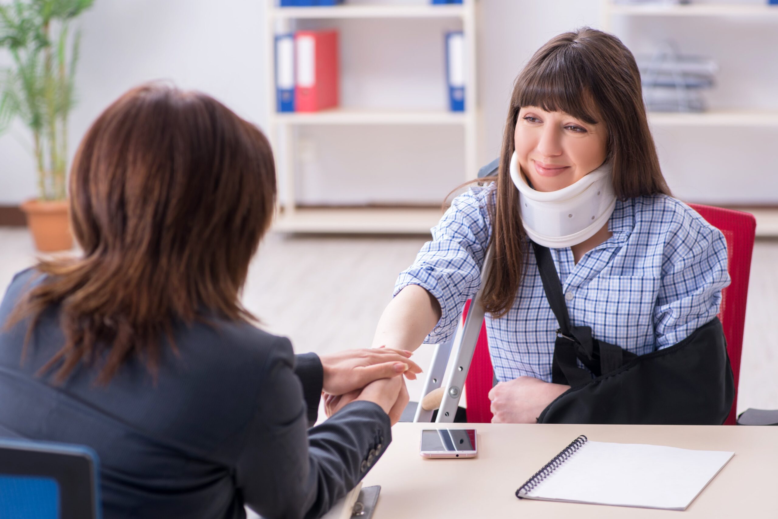 Injured woman with a neck brace and arm sling meeting with a female Car Accident Lawyer in a law office, holding hands across a desk during a supportive legal consultation.