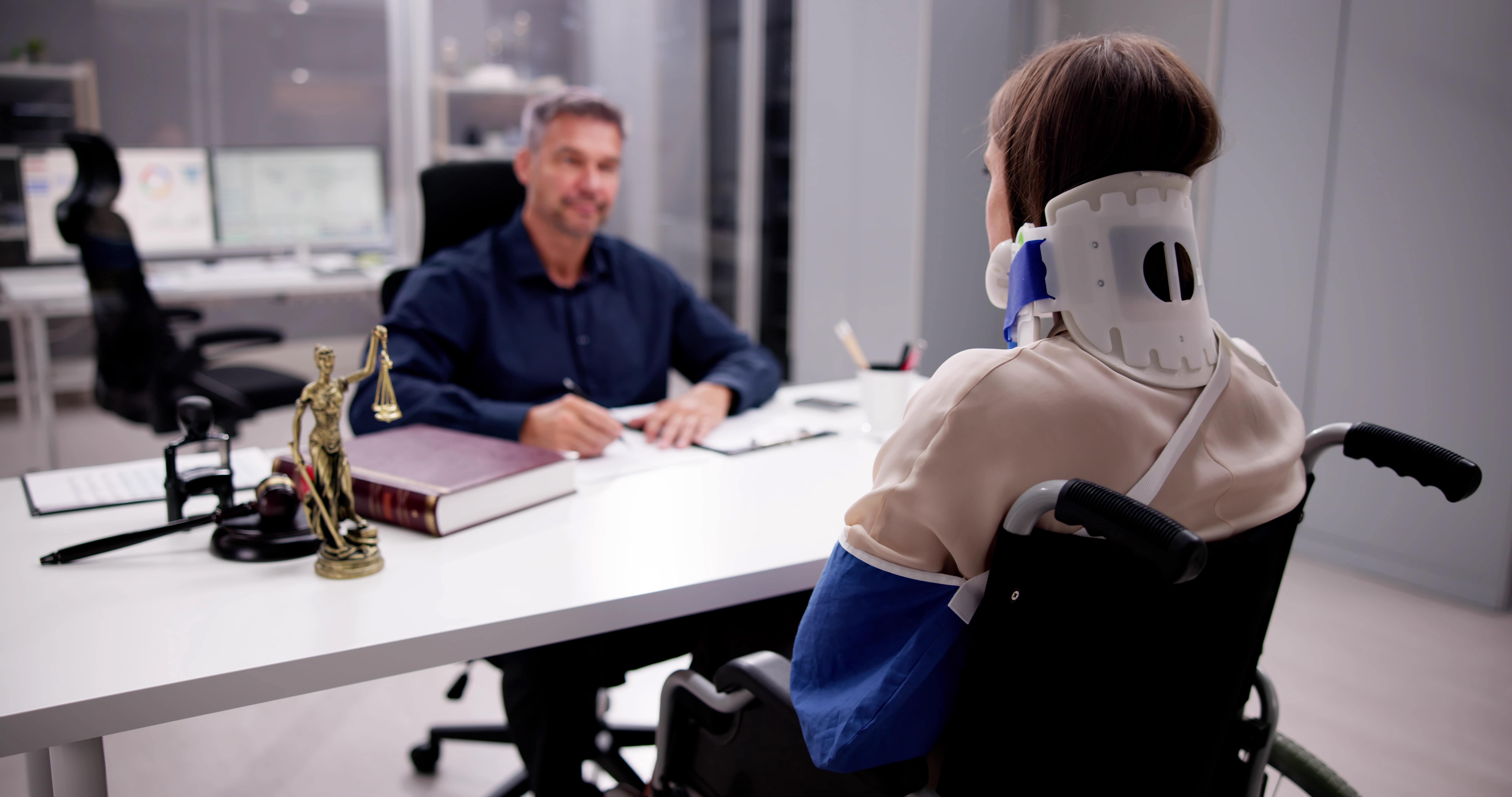 Injured woman in a wheelchair wearing a neck brace and arm sling consulting with a car accident lawyer in a modern legal office, discussing her case.